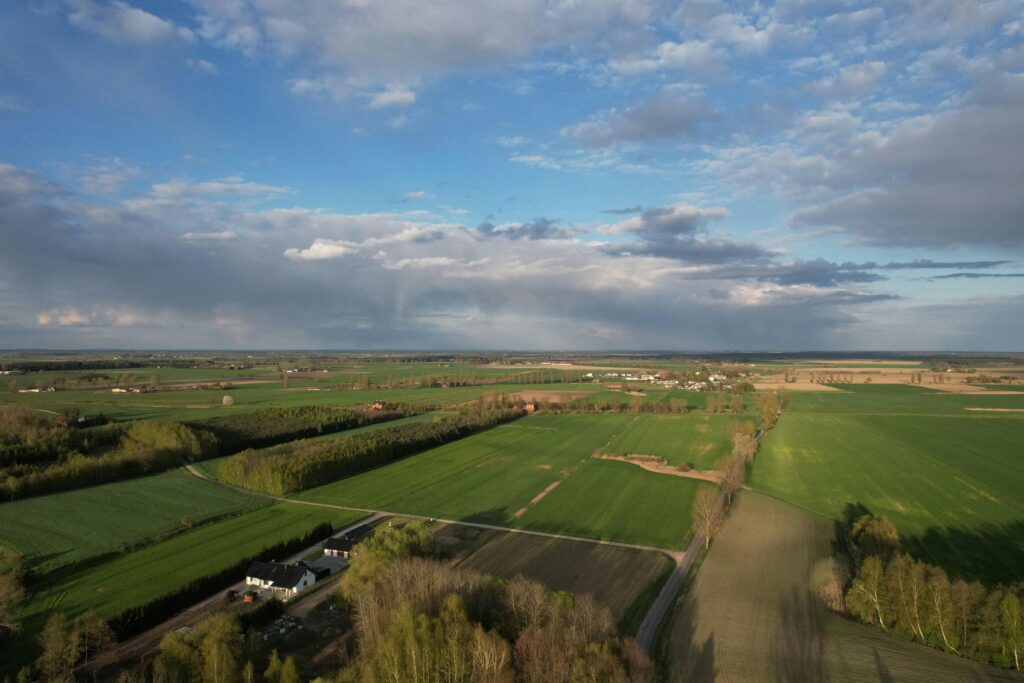 Aerial view of land with green fields, scattered trees under a partly cloudy sky.